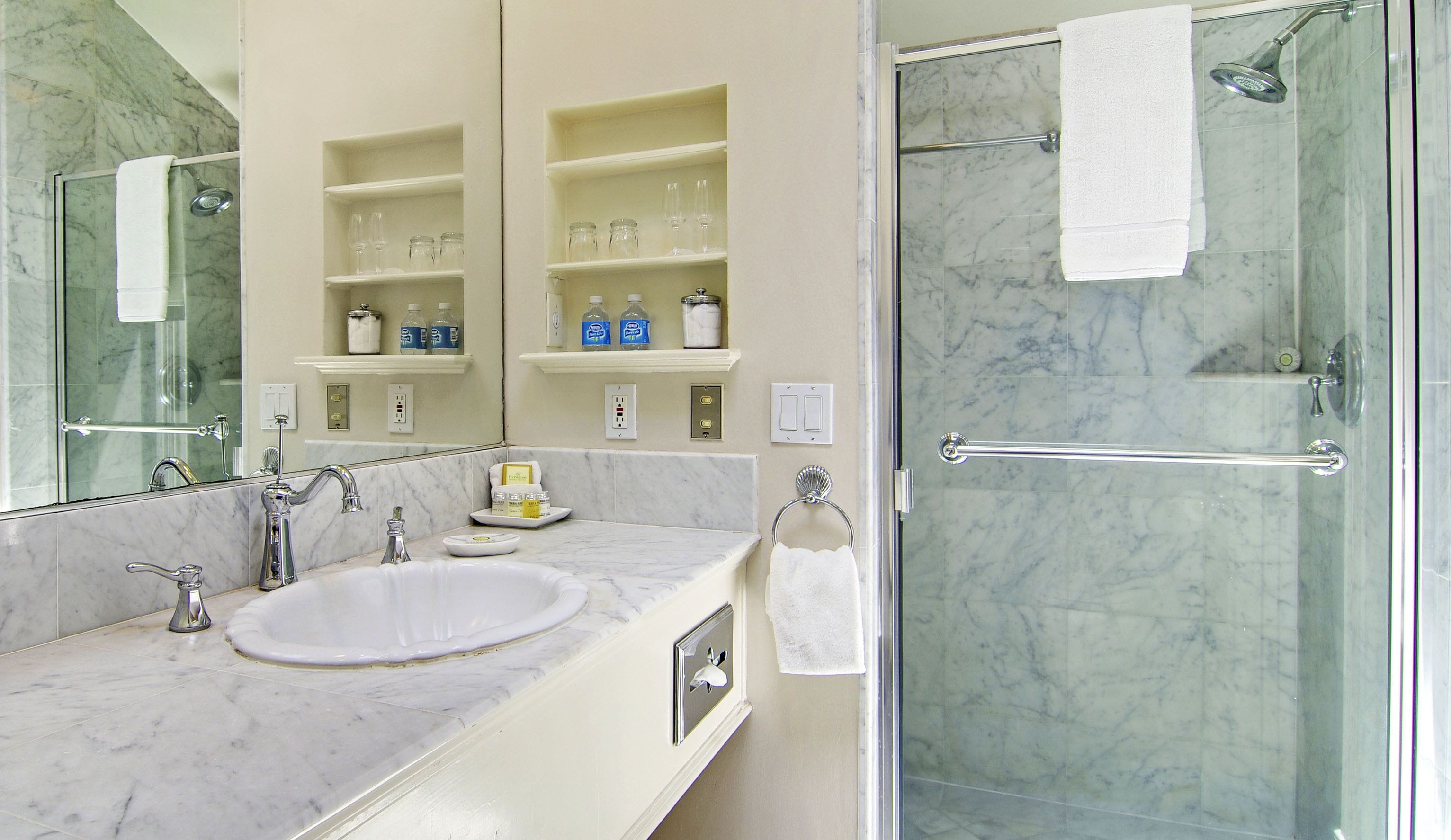 A bathroom with a marble sink, a glass-enclosed shower, and shelves with toiletries.
