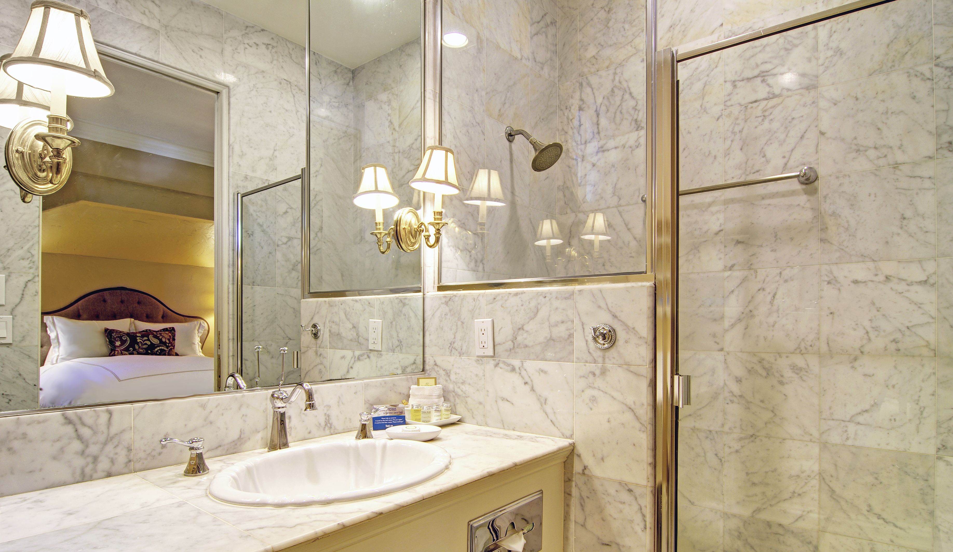 A bathroom with a large mirror, a sink, a shower, and a bed visible in the reflection.