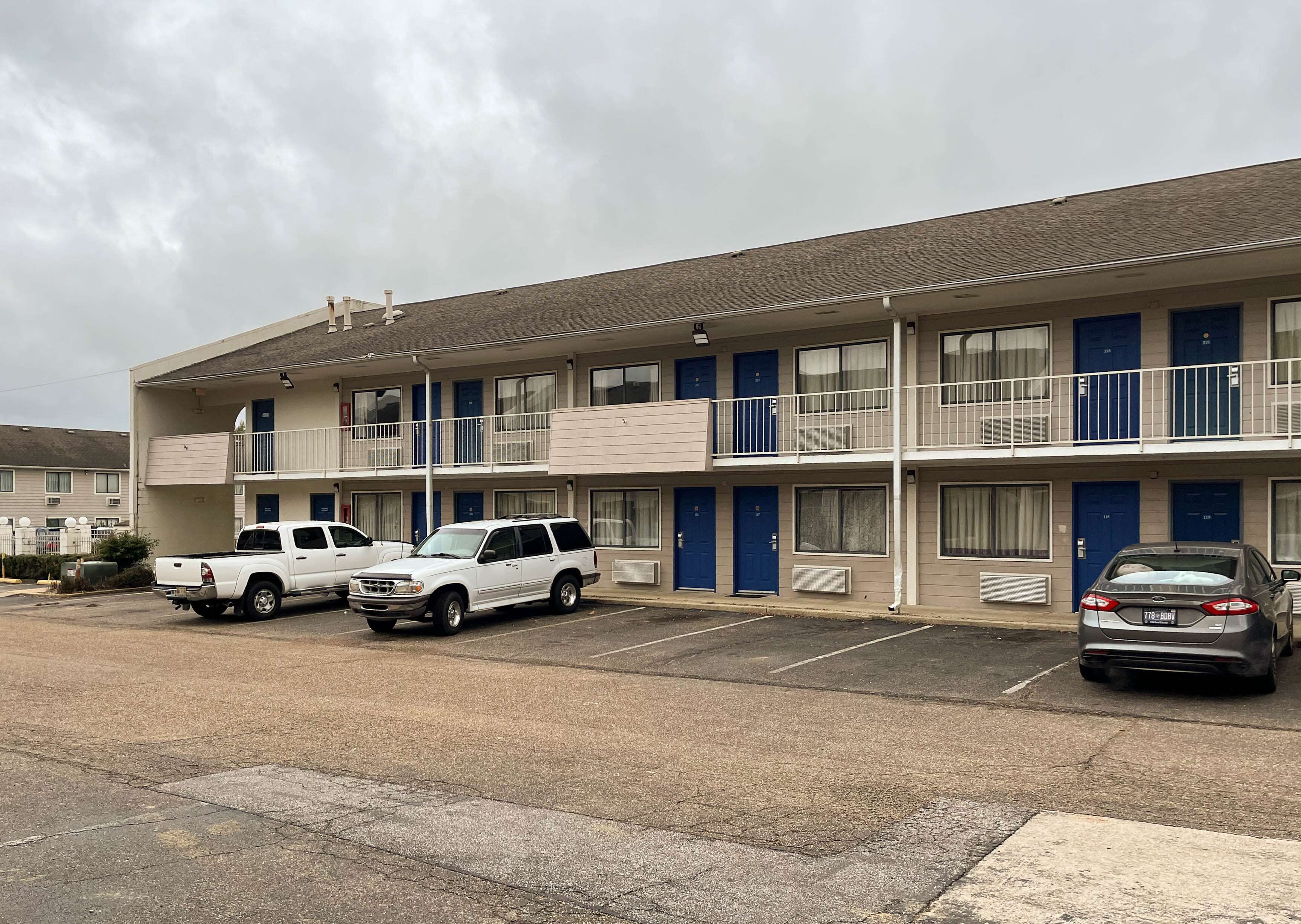 A motel with multiple floors, each with a balcony and a blue door. There are several parked vehicles, including a white pickup truck and a gray sedan.