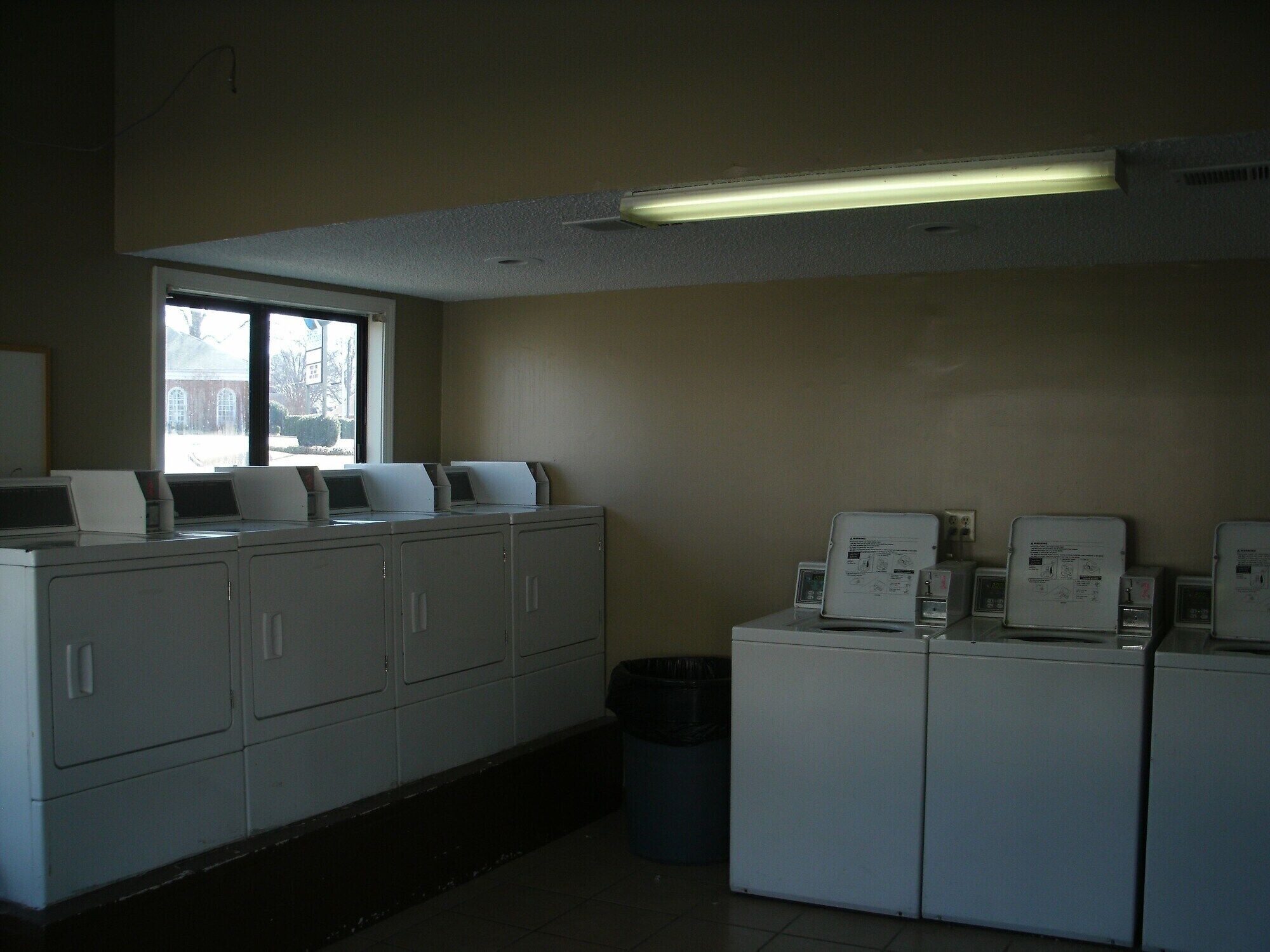 Une buanderie équipée de plusieurs machines à laver et de plusieurs sèche-linge, d’une poubelle et d’une fenêtre donnant sur un bâtiment à l’extérieur.