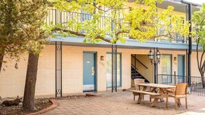 A building with a brick facade, a blue door, and a staircase leading to an upper level. There is a picnic table and chairs outside.