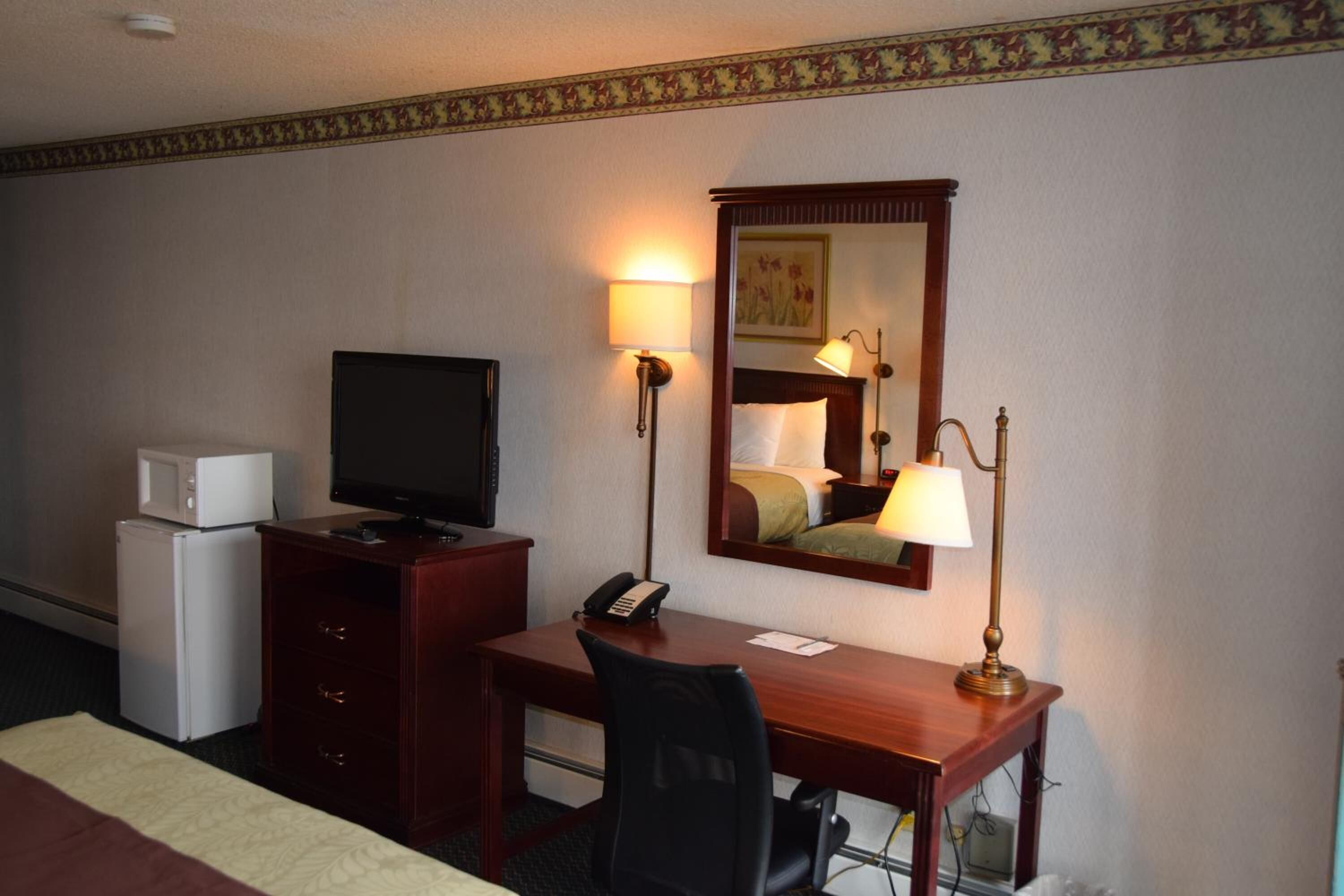 A hotel room with a wooden desk, a television, a telephone, a lamp, a mirror, a microwave, and a bed.