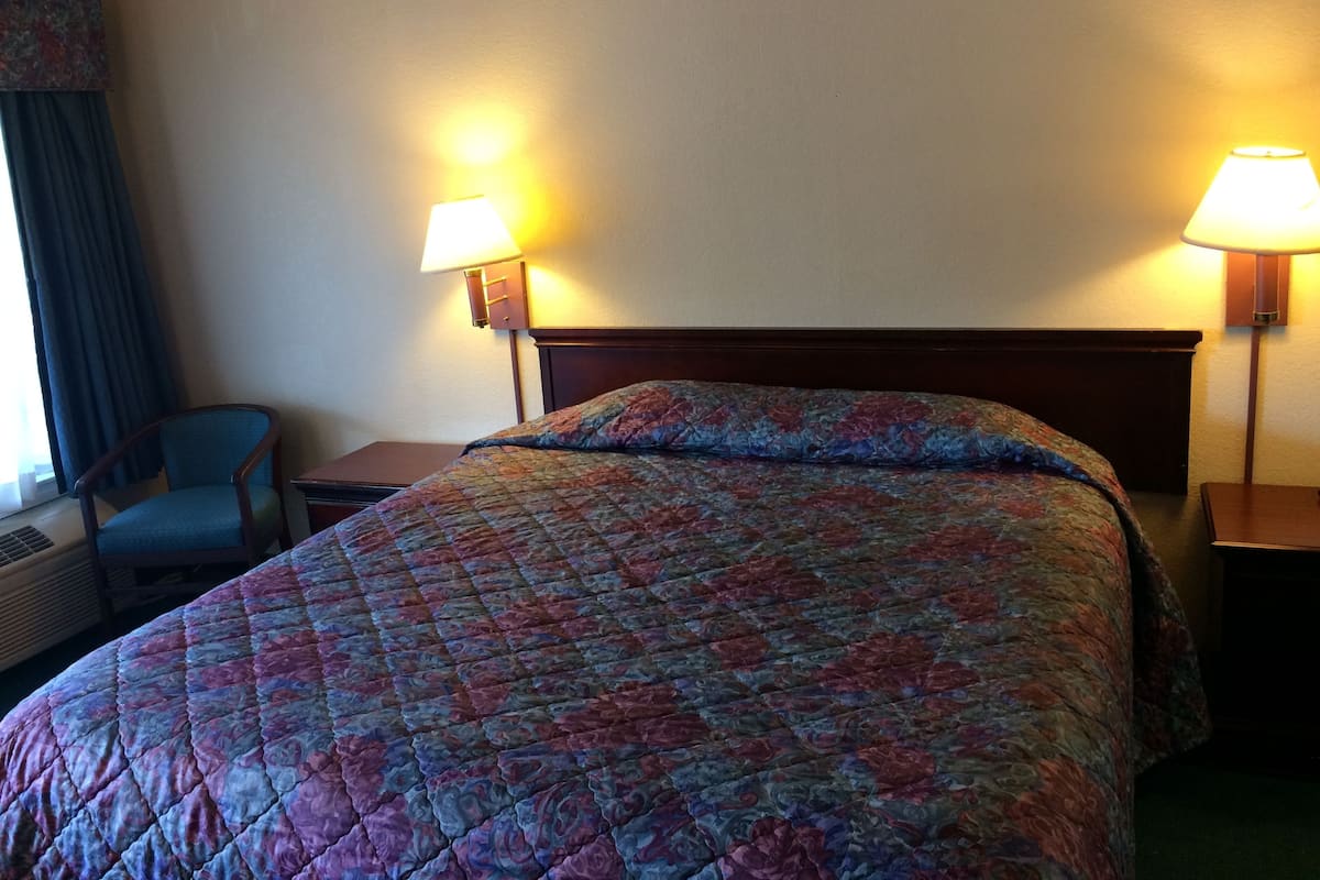Ein Hotelzimmer mit einem Bett, zwei Nachttischlampen, einem Sessel und einem Fenster mit Vorhängen.