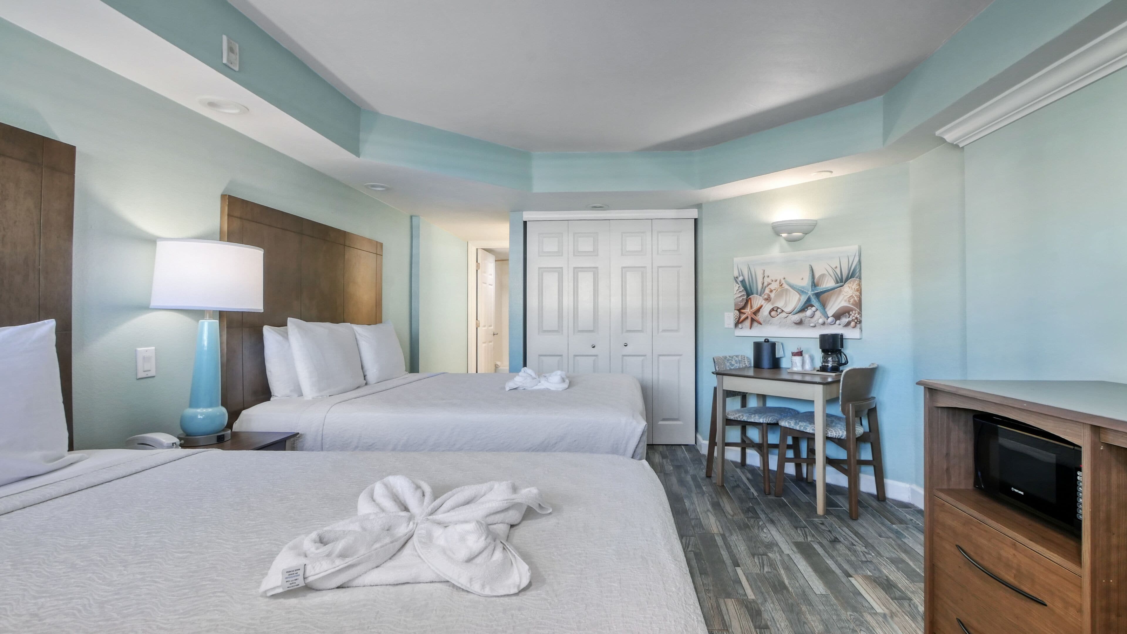 A hotel room with two beds, a desk with a chair, a coffee maker, and a painting on the wall.
