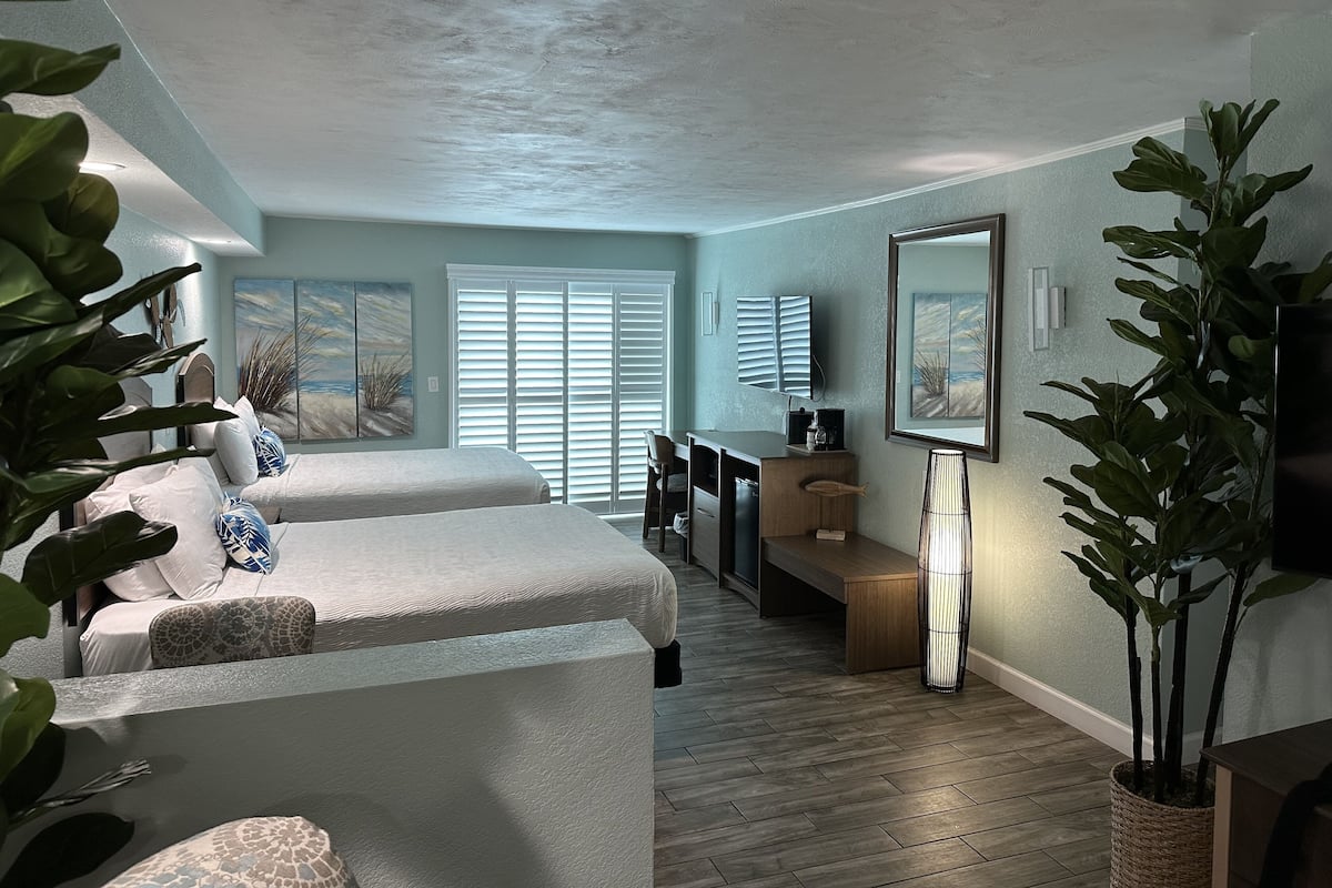 A bedroom with two beds, a desk, a chair, a lamp, and a large window with blinds.