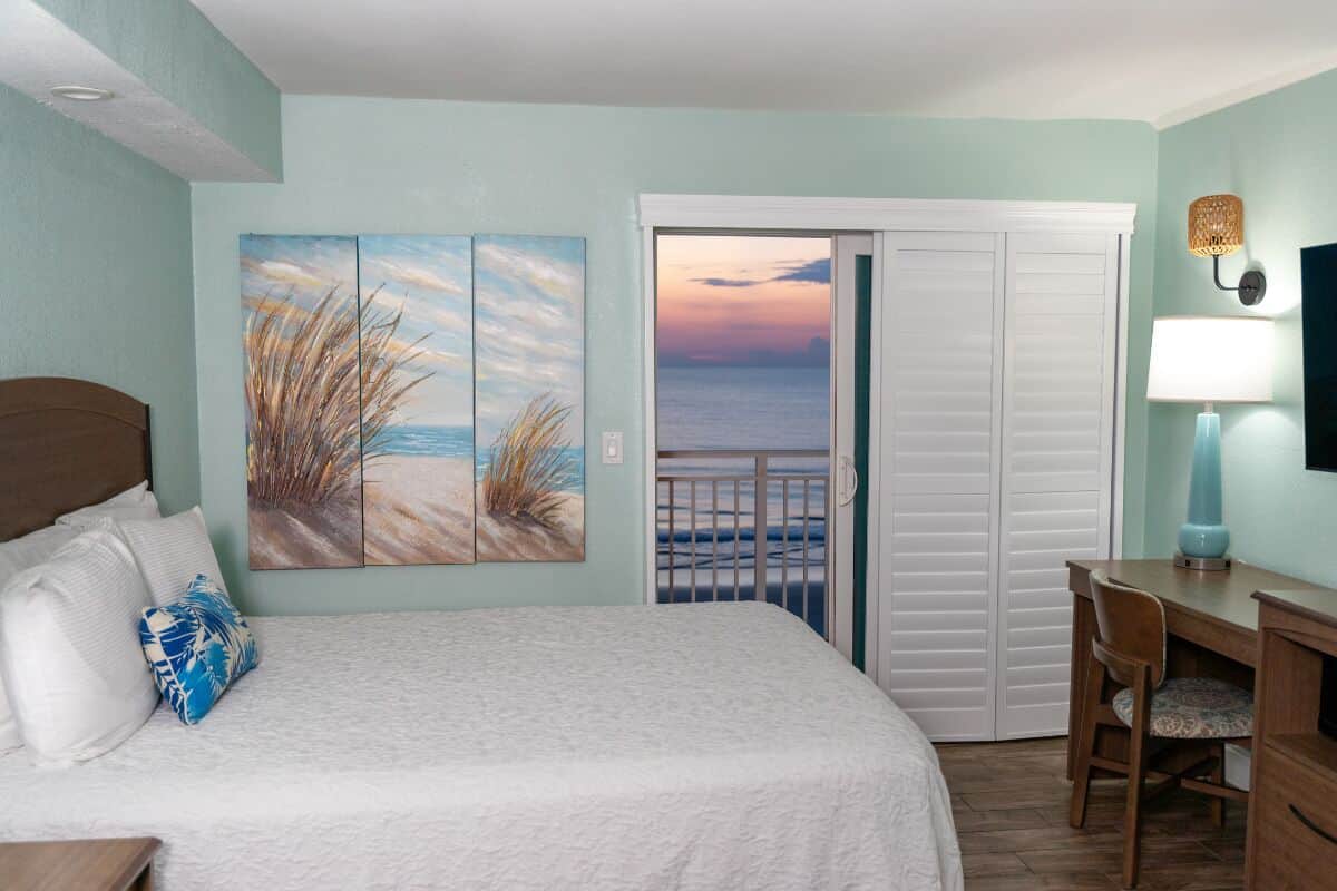 A bedroom with a bed, a desk, a chair, a painting of a beach, and a view of the ocean through a door.