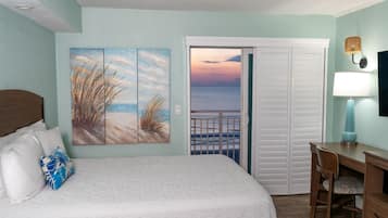 Un dormitorio con cama, escritorio, silla, un cuadro de playa y vista al océano a través de una puerta.