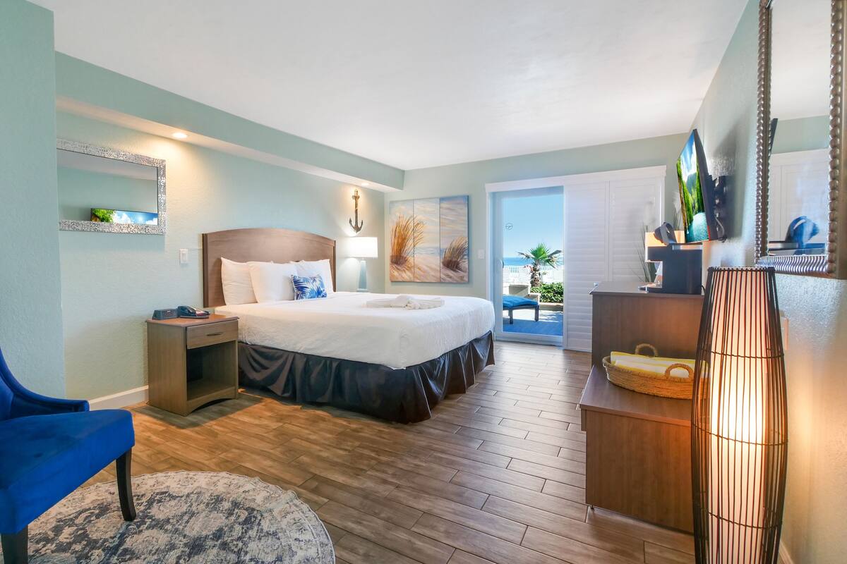 A hotel room with a bed, a blue chair, a nightstand, a lamp, a painting, a view of the beach, and a wooden floor.