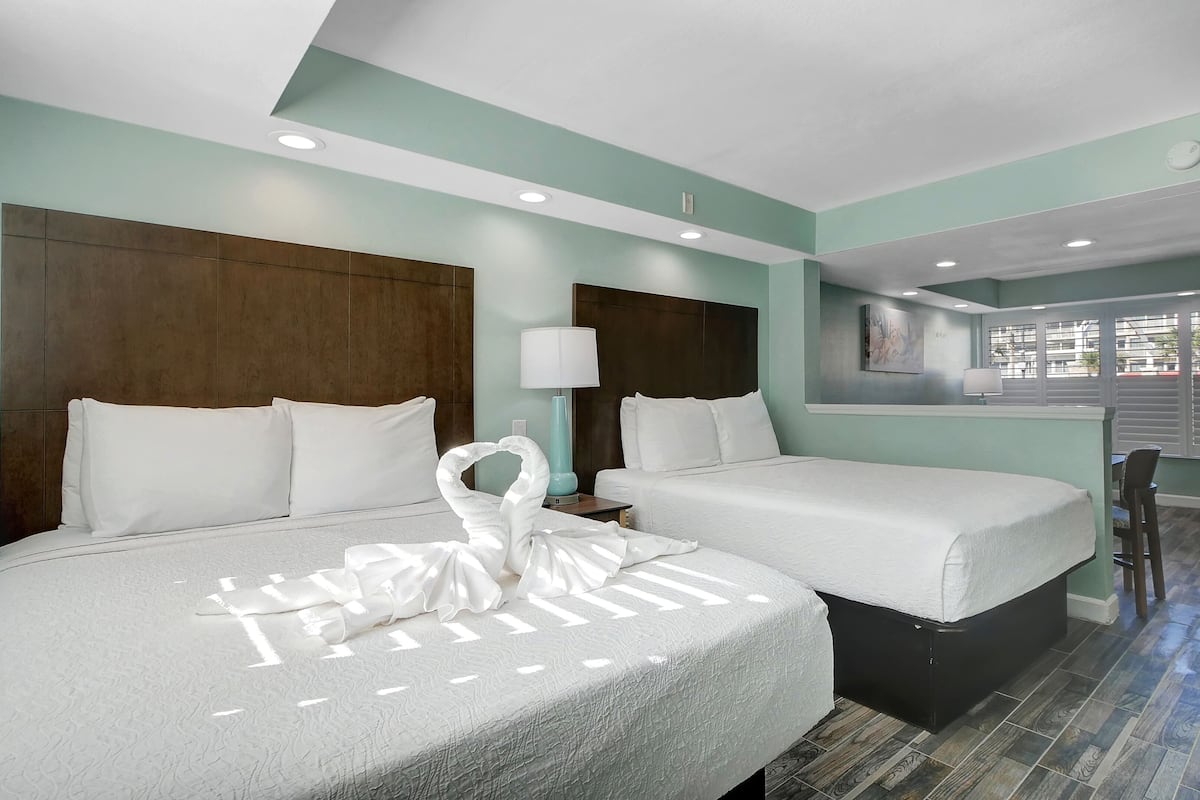 A hotel room with two beds, a small table, a chair, and a window with blinds.