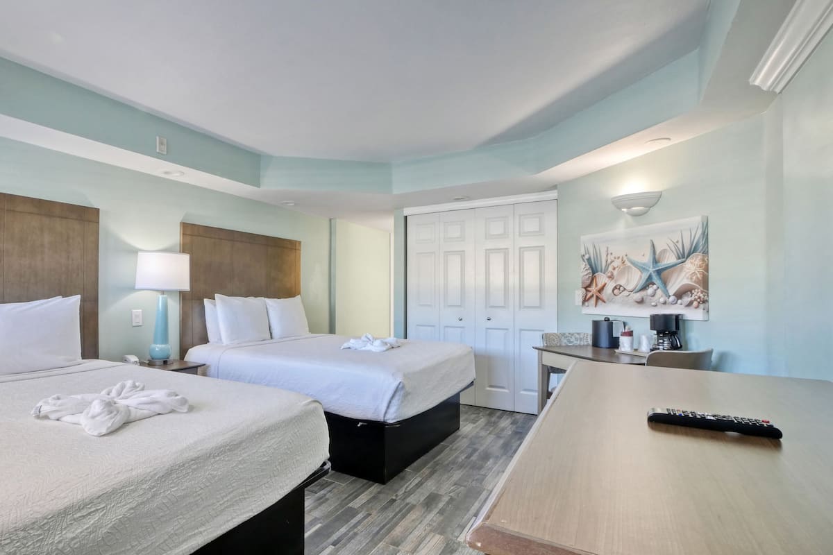 A hotel room with two beds, a desk, a chair, a lamp, a painting, and a remote control.