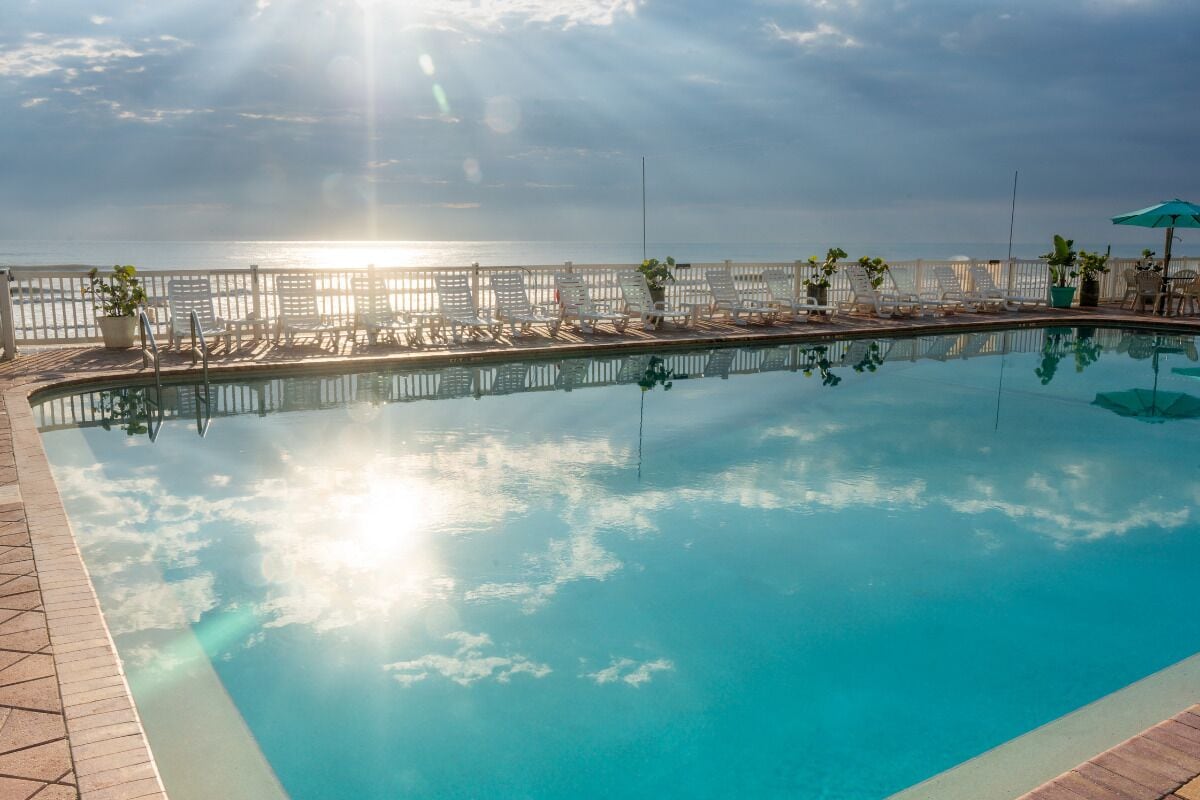 A clear swimming pool with sun reflecting on the water, surrounded by lounge chairs and umbrellas, with a view of the sea in the background.