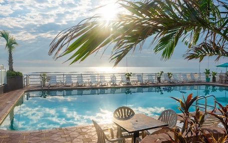 A pool area with a view of the sea, lounge chairs, and a table with chairs.