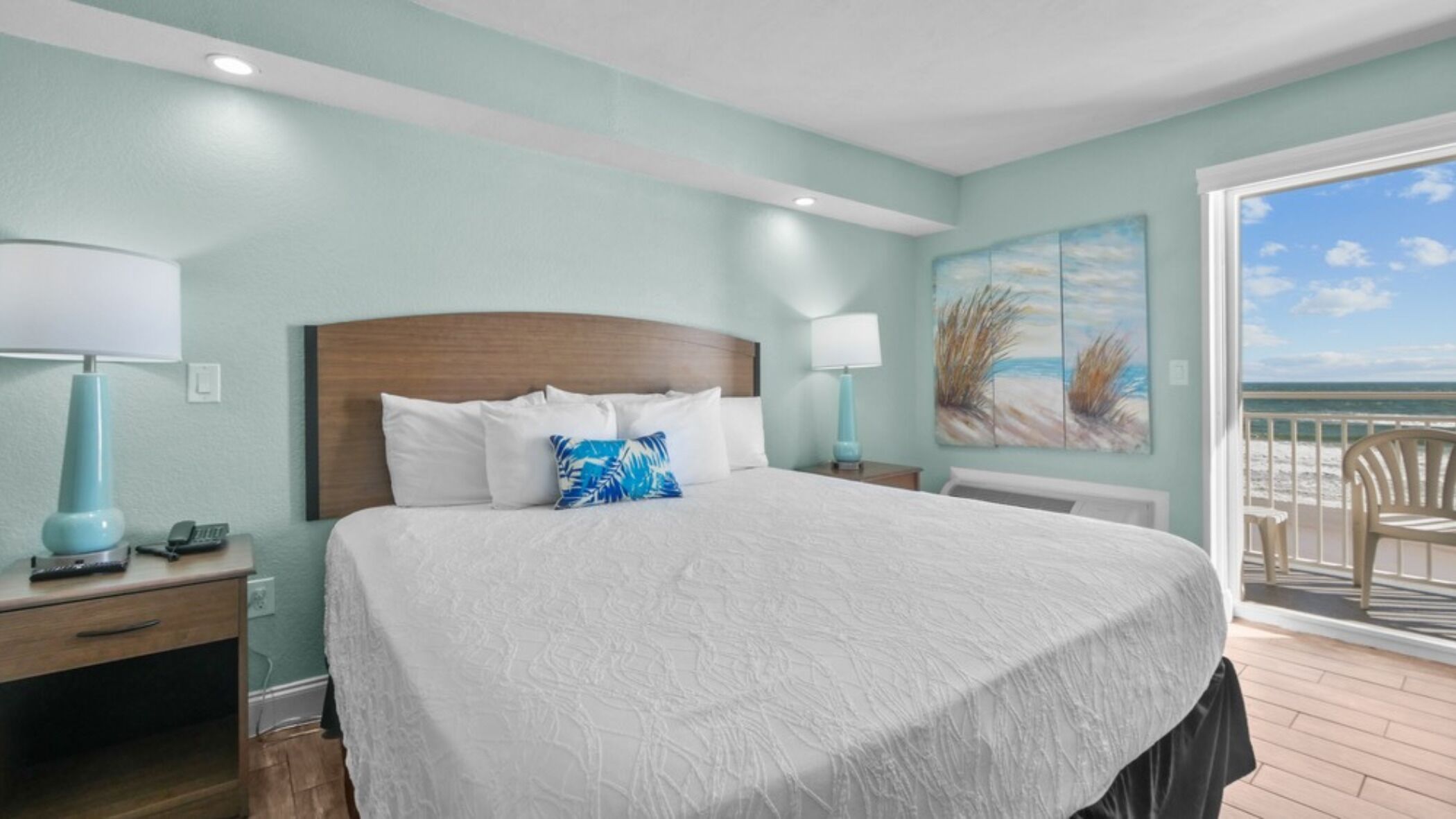 A bedroom with a large bed, two bedside tables, a nightstand, a window with a view of the beach, and a painting on the wall.