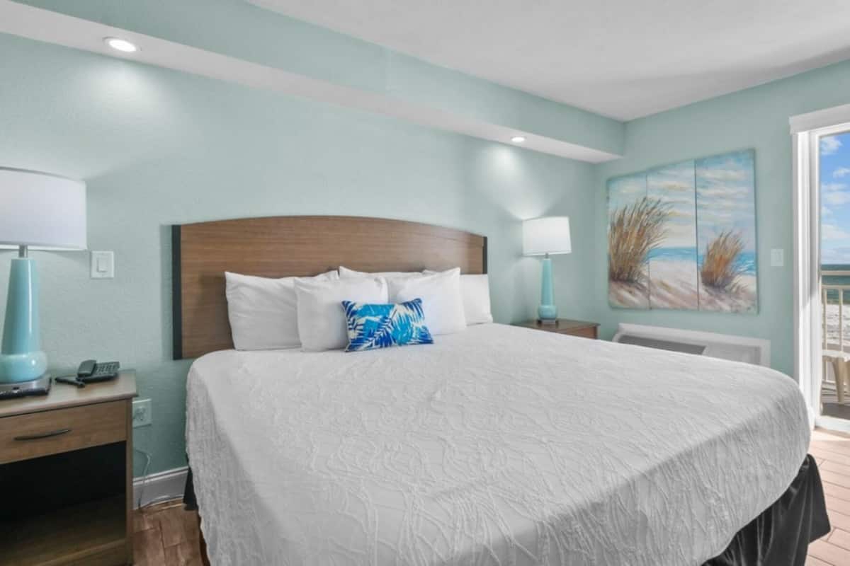 A bedroom with a large bed, two bedside tables, a nightstand, a window with a view of the beach, and a painting on the wall.