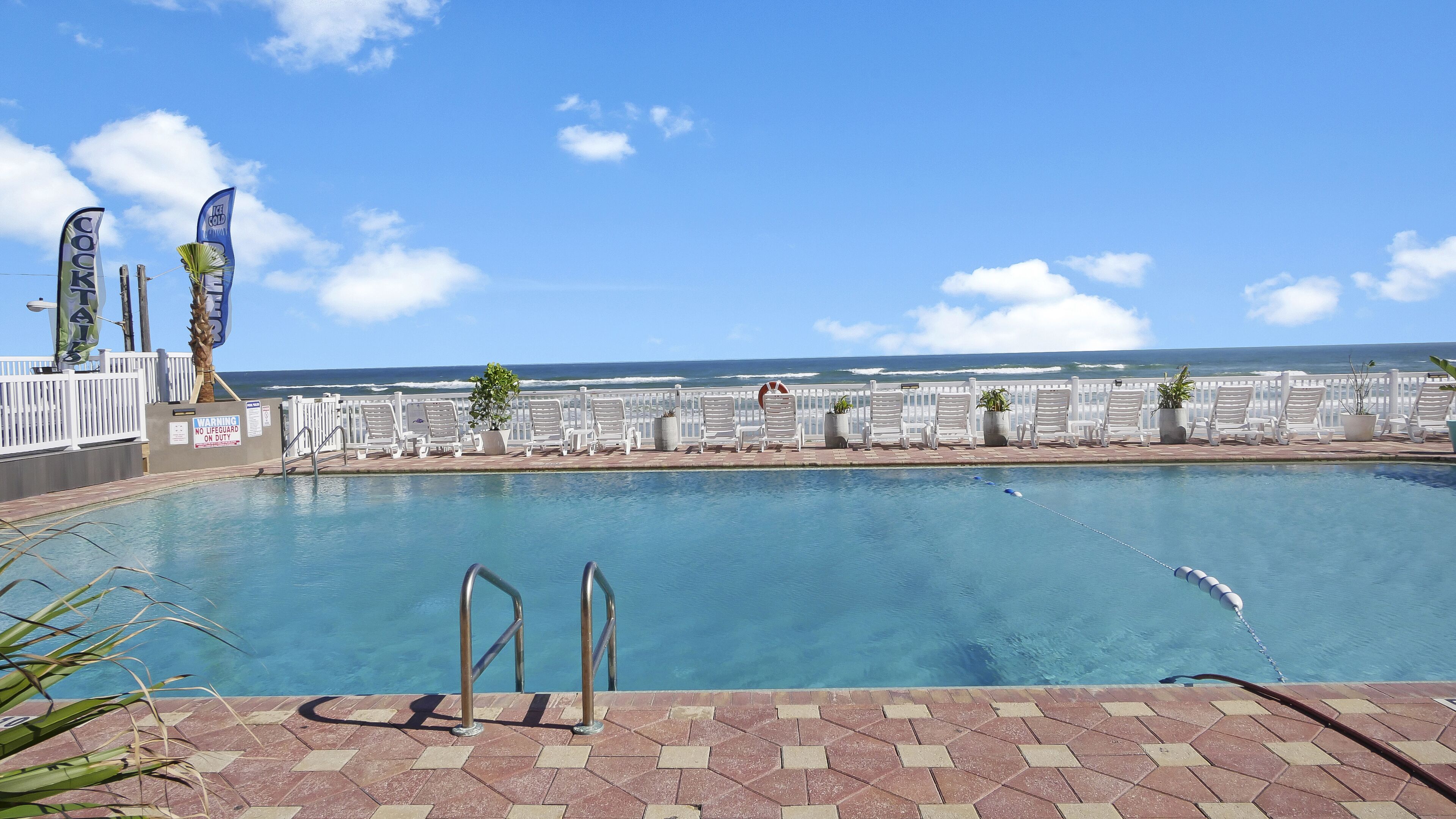 A pool area with a view of the ocean, white lounge chairs, and flags.