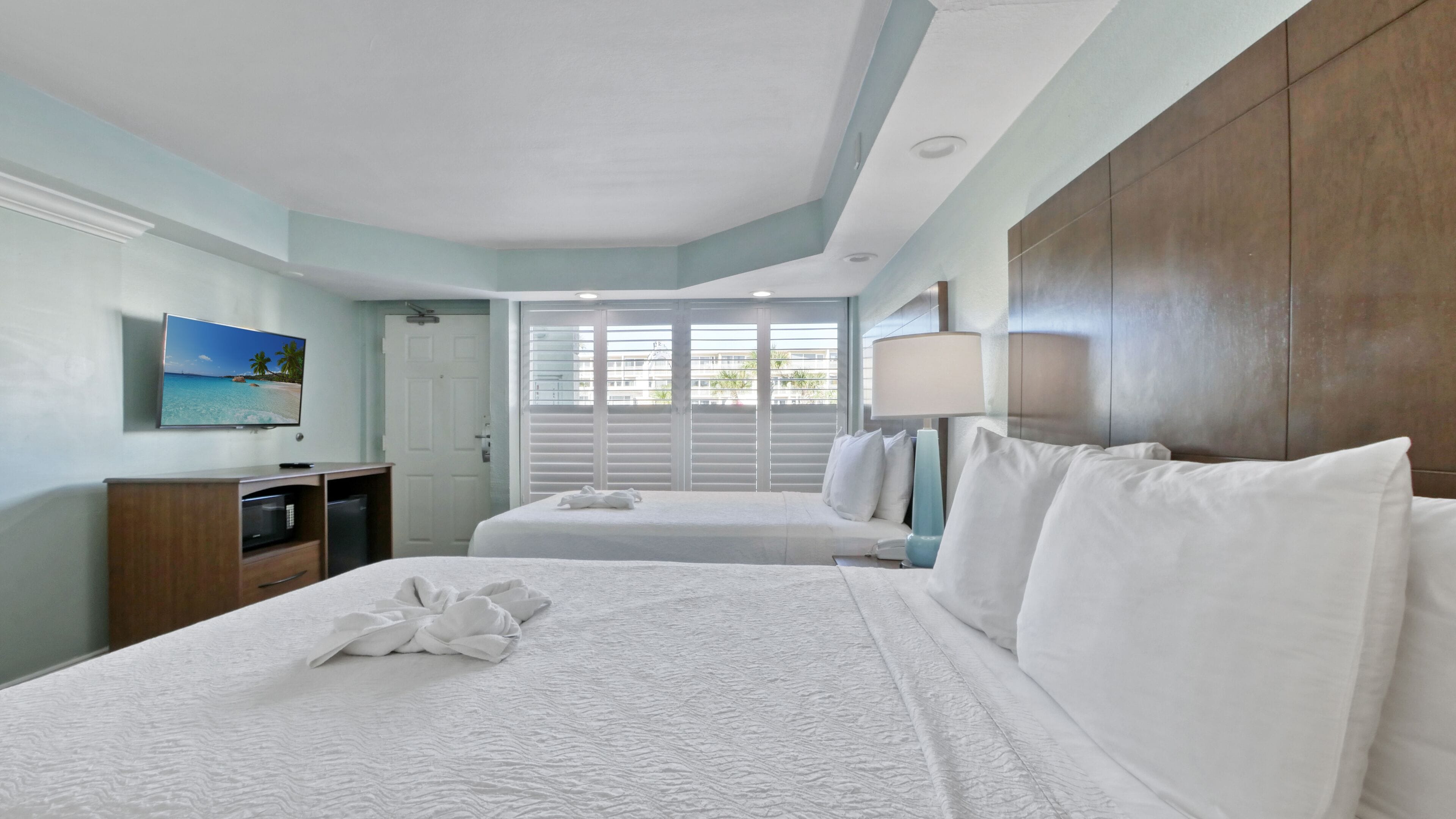 A hotel room with two beds, a TV, a desk, and a window with blinds.
