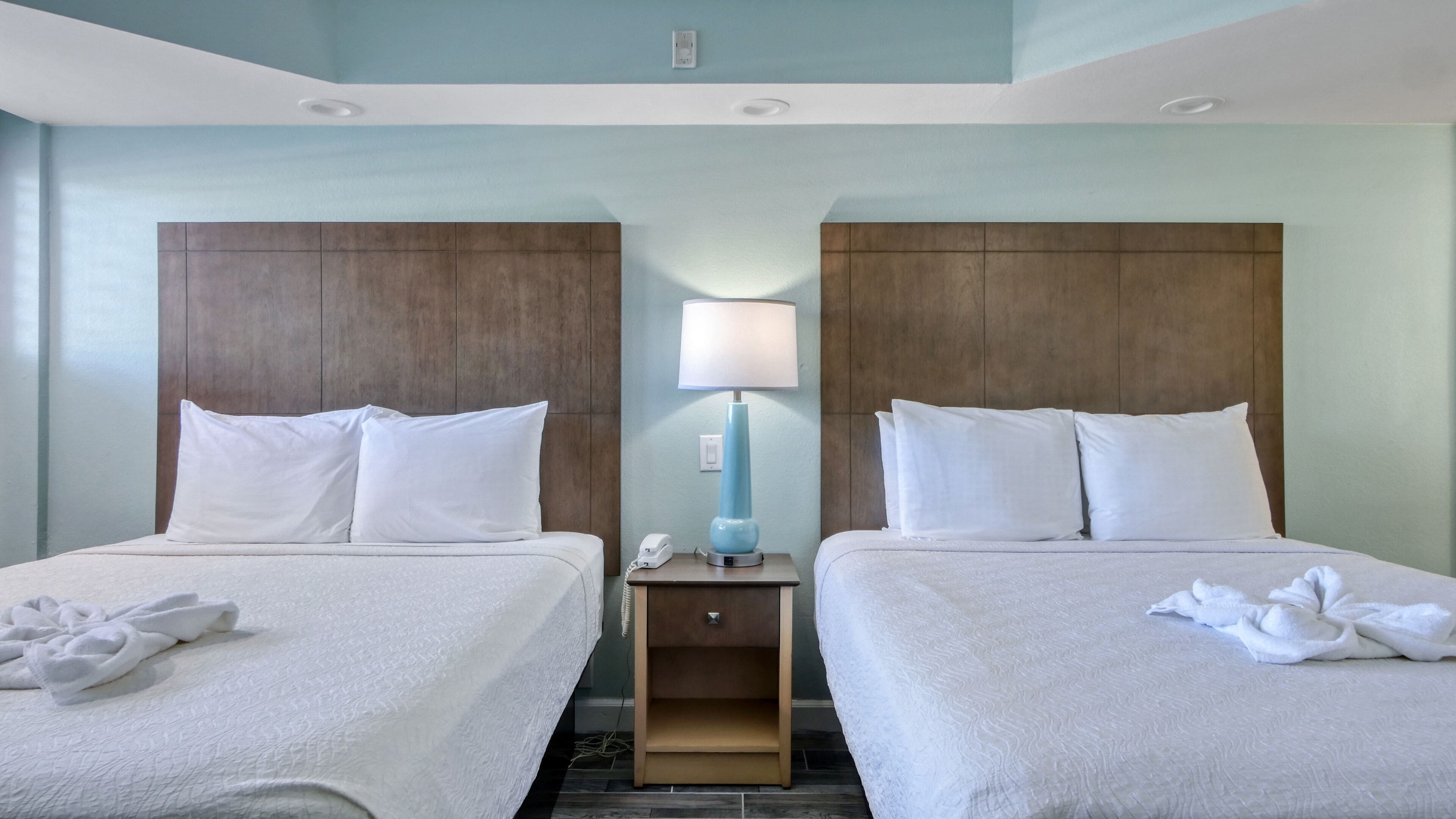 Habitación de hotel con dos camas, cada una con ropa de cama y almohadas blancas, una mesita de noche con lámpara y teléfono.