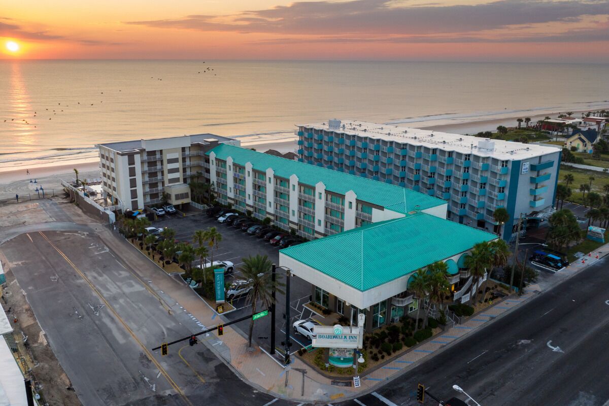 Un hotel con un edificio principal y una sección más pequeña, ambos con techo verde, ubicado frente a una playa y con vista despejada al océano.