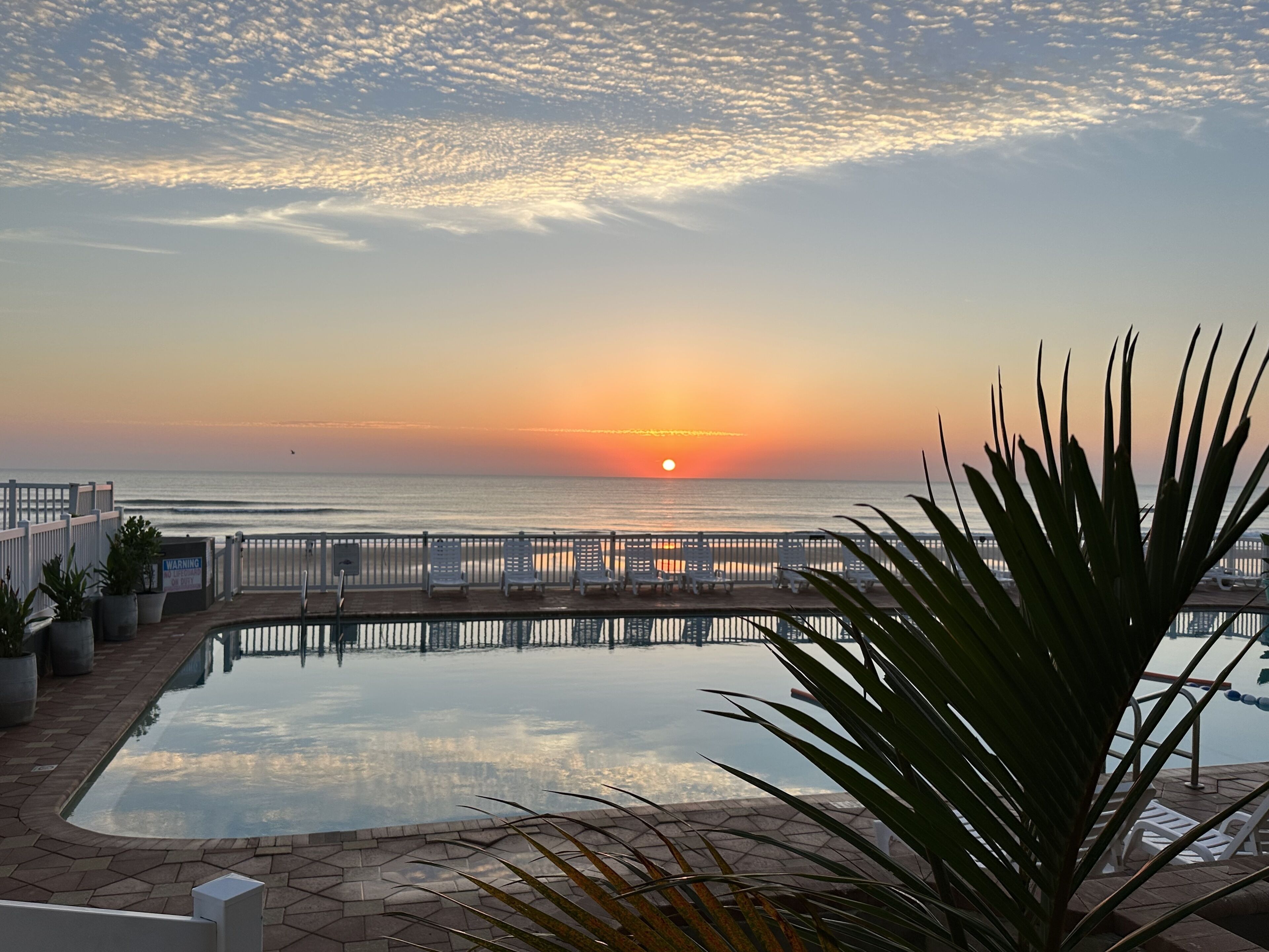 A sunset view from a poolside area with a fence and lounge chairs.