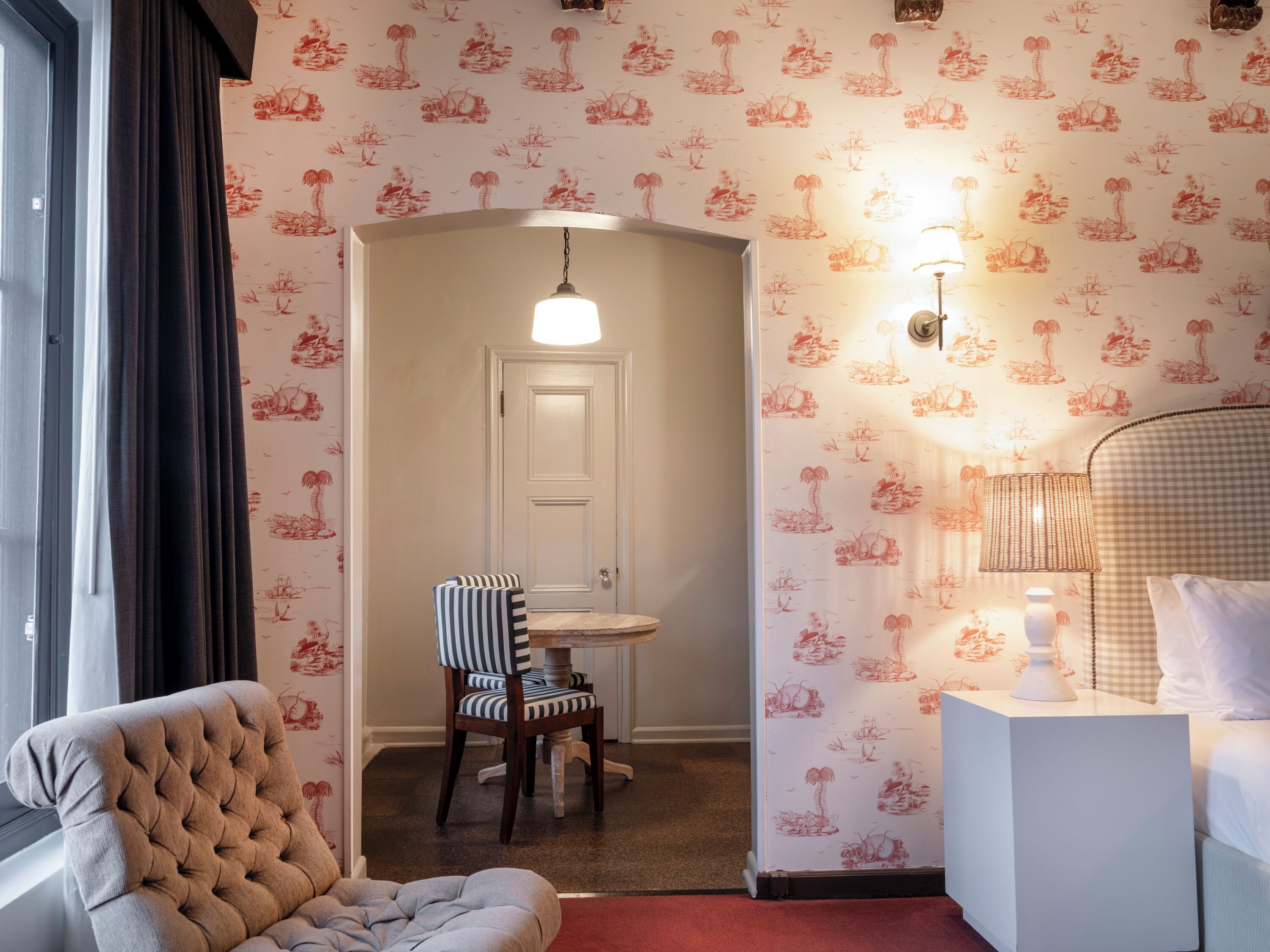 A hotel room with a checkered headboard, a tufted armchair, a striped chair, a small round table, and a wall with a red and white patterned wallpaper.