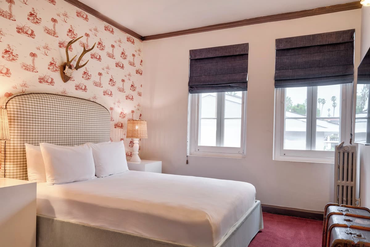 A bedroom with a checkered headboard, a white bed, two bedside lamps, a radiator, a window with a view, and a suitcase.