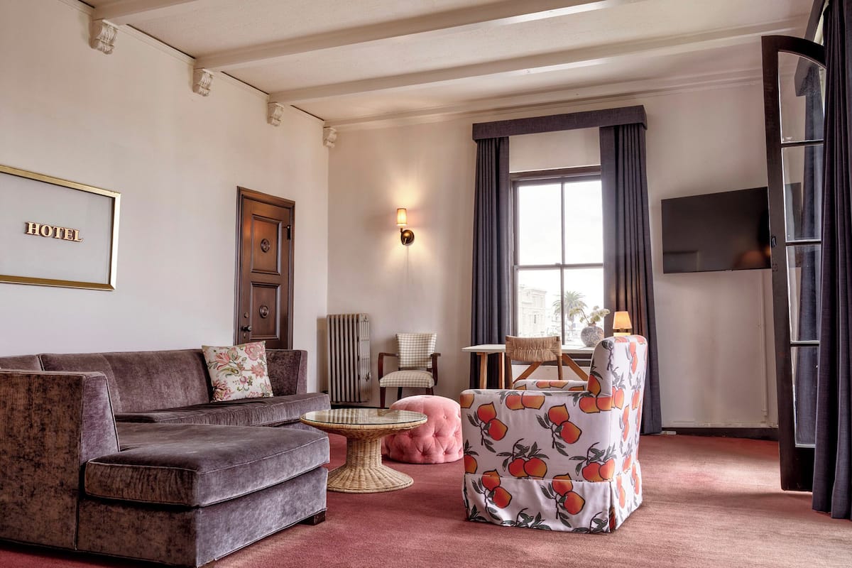 A hotel room with a grey sofa, a round wicker table, a pink ottoman, a chair with a fruit pattern, a wall-mounted TV, and a large window with curtains.