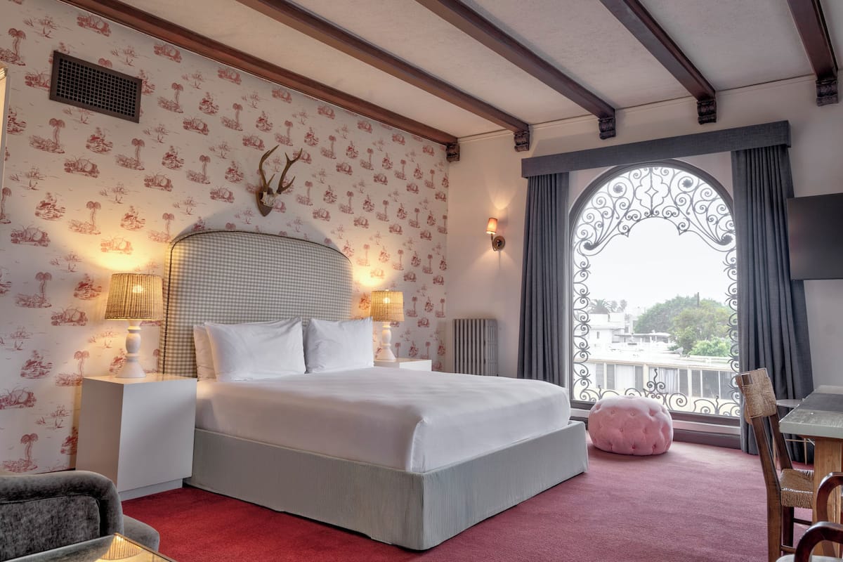 A hotel room with a large bed, two bedside lamps, a decorative wall with a patterned wallpaper, a ceiling with exposed beams, and a balcony with a wrought iron gate.