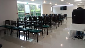 Une salle de conférence avec des rangées de chaises, une grande fenêtre donnant sur un paysage verdoyant et un téléviseur à écran plat fixé au mur.