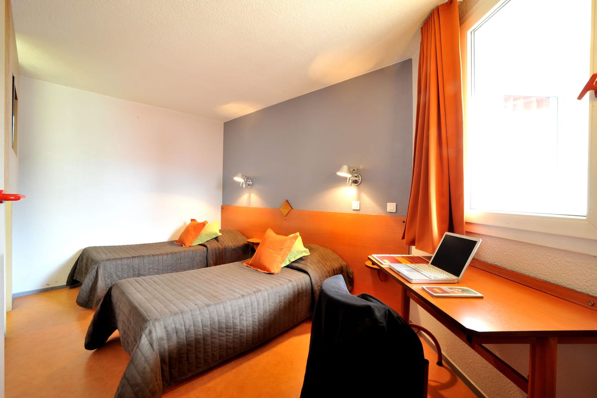 A hotel room with two beds, a desk with a laptop, and a window with curtains.