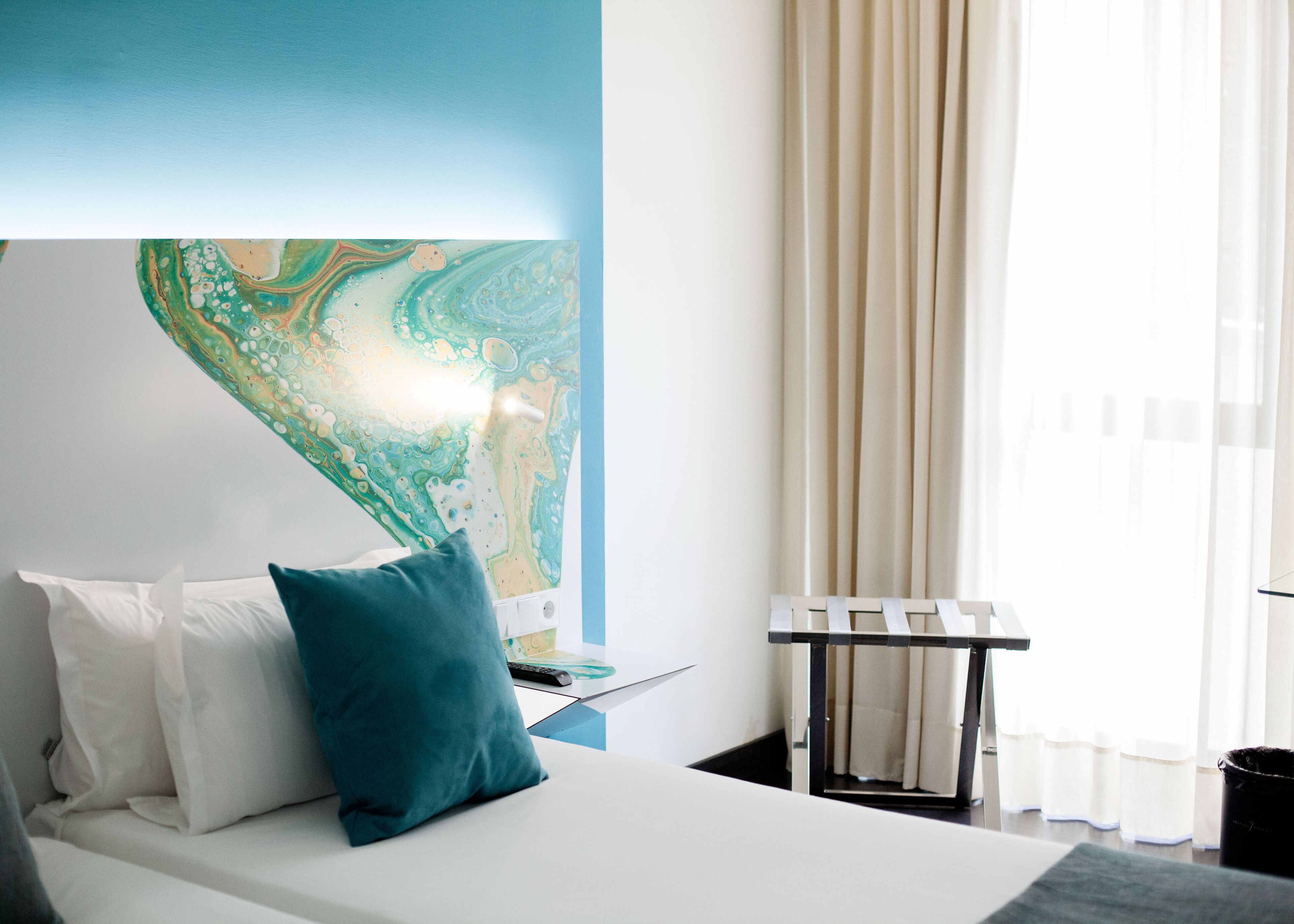 Una habitación de hotel moderna con un gran mural abstracto en la pared, una cama con almohadas blancas y turquesa, y una ventana con cortinas translúcidas.