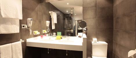 A modern bathroom with a large mirror, white sink, and a toilet. There are towels hanging on the wall and a trash can in the corner.