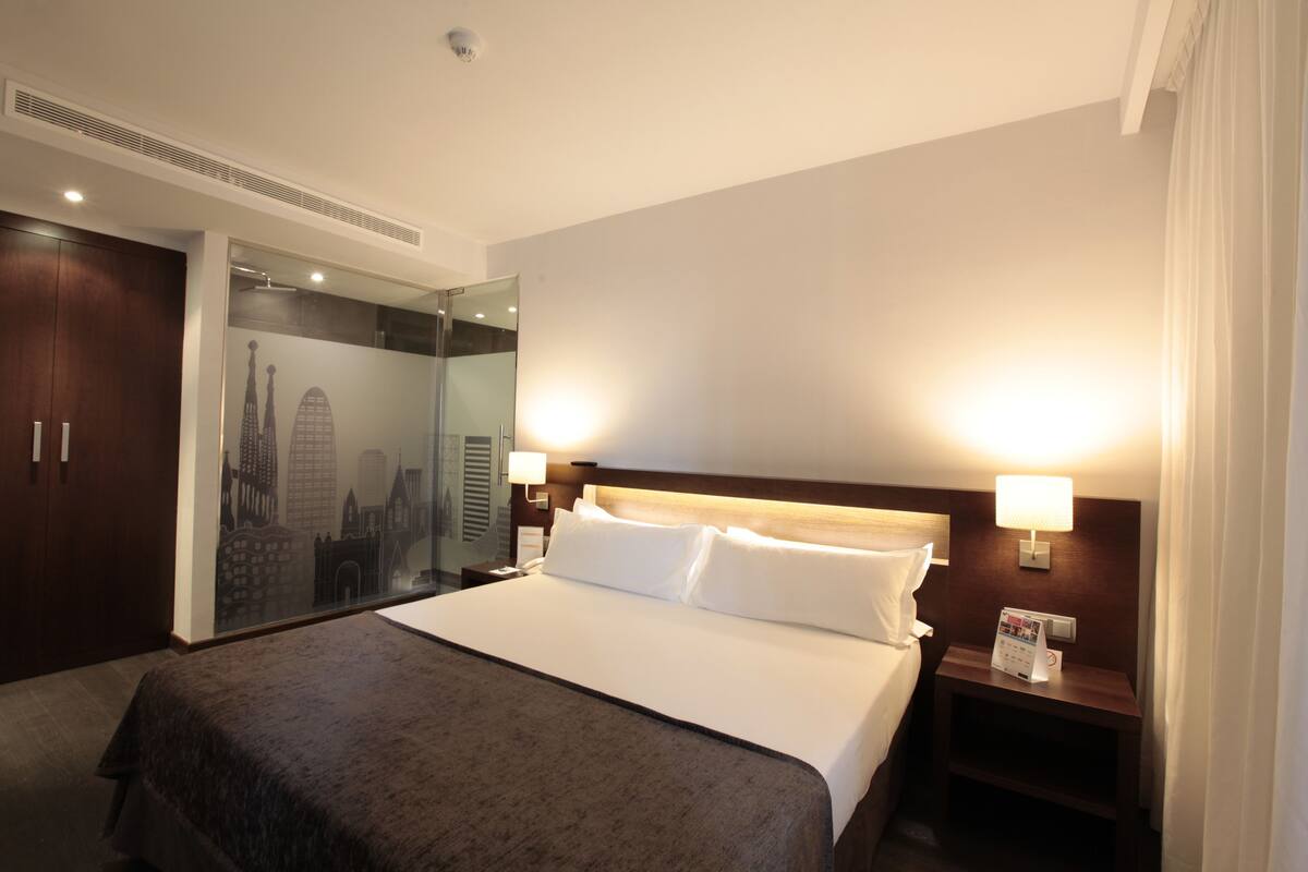 A hotel room with a large bed, a wooden headboard, two bedside lamps, a nightstand with a clock and a magazine rack, a wardrobe, and a mirror with a cityscape design.