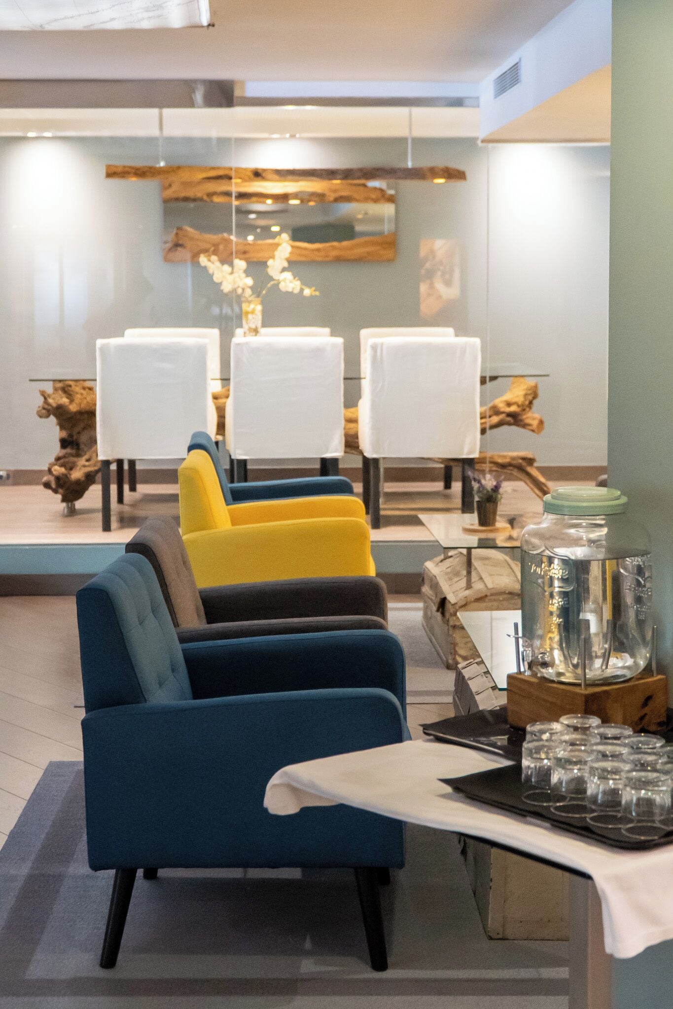 A modern lounge area with blue and yellow armchairs, white tables, and a decorative wooden piece on the wall.
