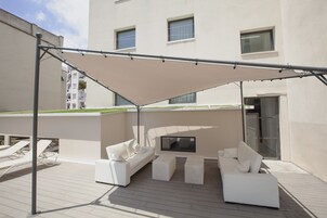 Una terraza en la azotea con mobiliario exterior blanco, un toldo grande y vistas de un edificio al fondo.
