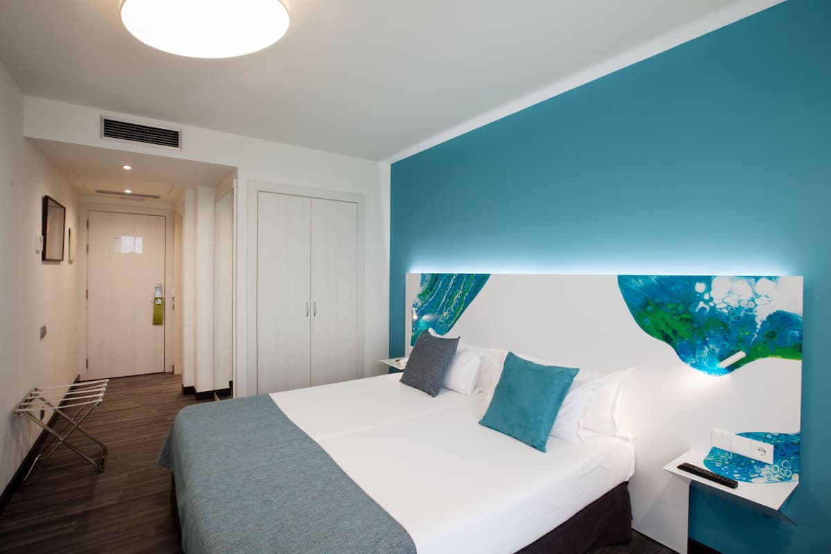 A modern hotel room with a large bed, a nightstand, a wall art piece, and a door leading to another room.