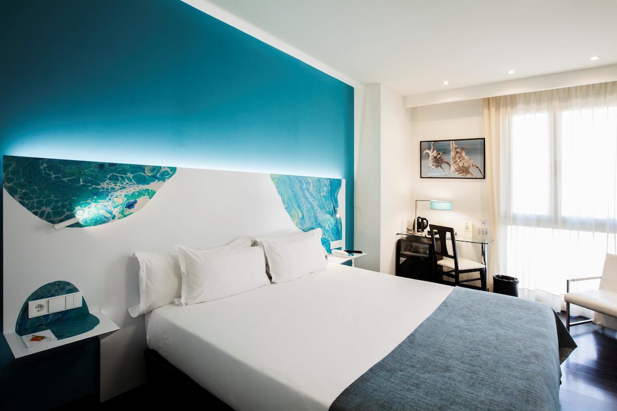 A hotel room with a large bed, a desk with a chair, a painting on the wall, and a window with curtains.