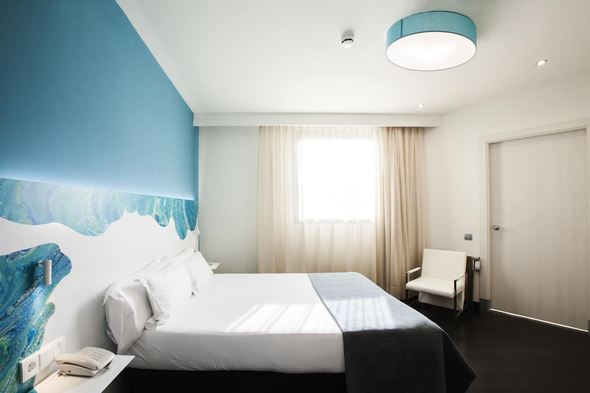 A hotel room with a bed, a chair, a desk with a phone, a window with curtains, and a wall with a blue abstract design.
