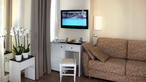 Une chambre d’hôtel équipée d’un bureau, d’un téléviseur fixé au mur, d’un canapé et d’un petit tabouret blanc.