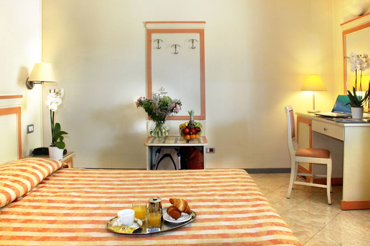 Een hotelkamer met een bed, een bureau, een stoel, een klein tafeltje met een schaal fruit, een vaas met bloemen en een dienblad met ontbijtartikelen.
