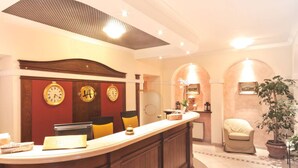 A reception area with a wooden counter, two clocks, a plant, and a seating area with a chair. - Hotel Villa Ambrosina (Impruneta)