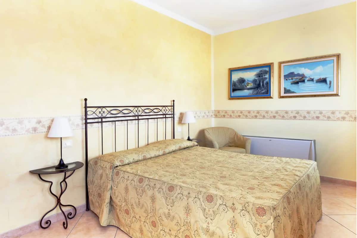 A bedroom with a bed, two wall-mounted paintings, a bedside table with a lamp, and a chair.