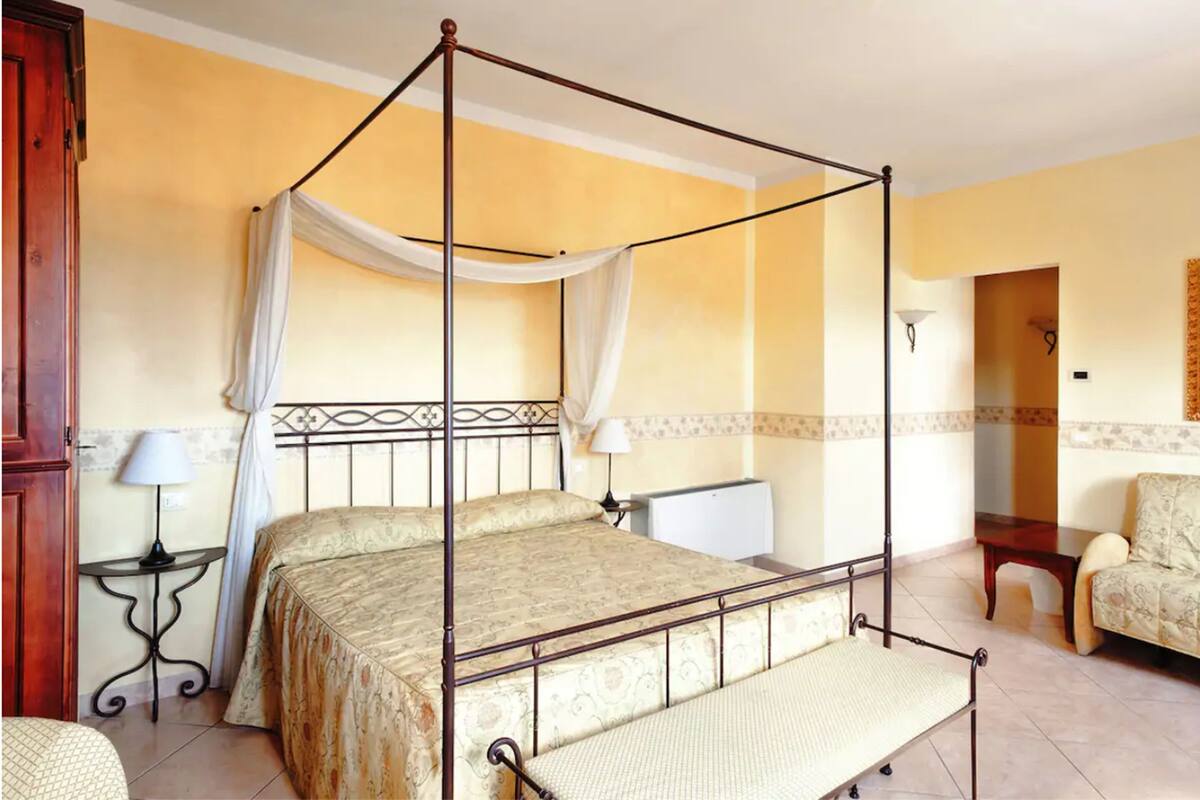 A four-poster bed with a canopy, a bedside table with a lamp, a chair, and a small table with a lamp.