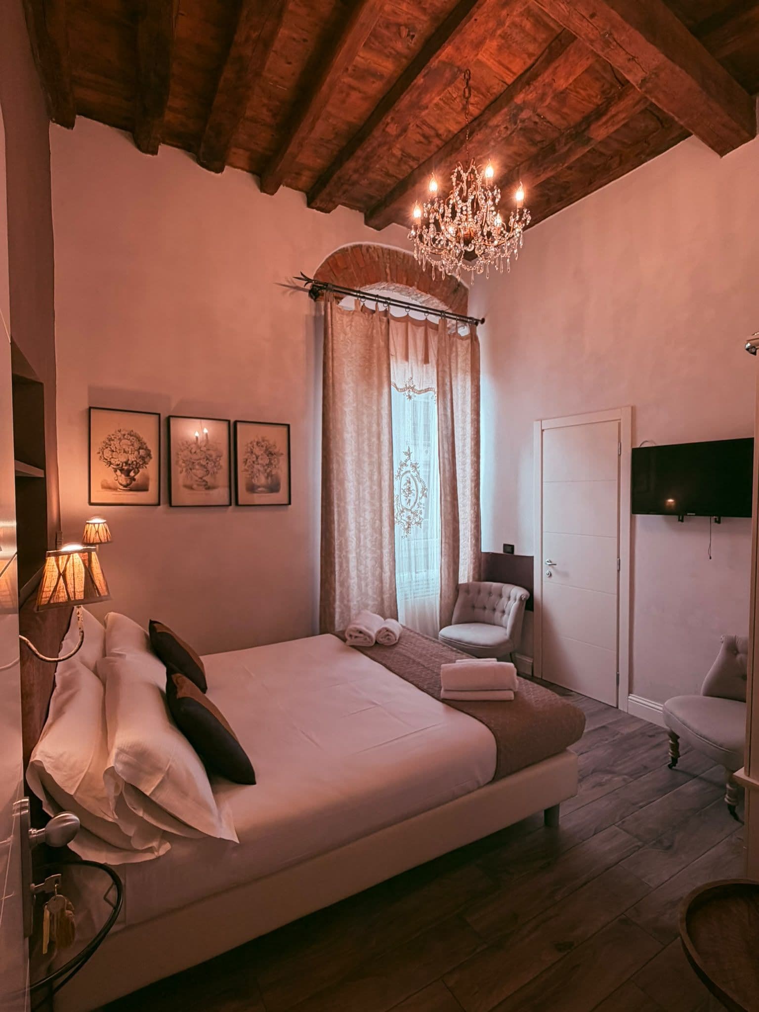 A bedroom with a large bed, a chandelier, a chair, a television, and artwork on the wall.