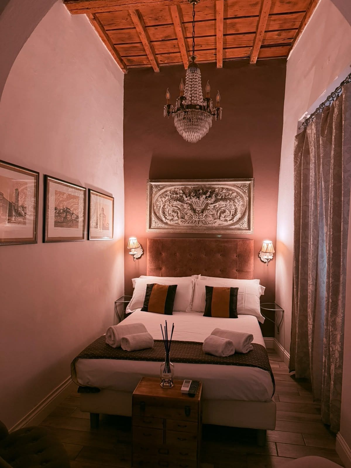 A hotel room with a large bed, a chandelier, framed pictures on the wall, and a wooden headboard.