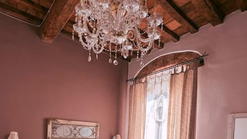 Un dormitorio con una cama grande, dos sillones, una mesita redonda, una araña y un ventanal con cortinas.