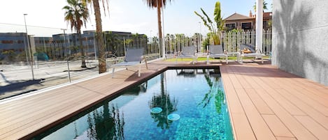 A swimming pool with a tiled deck, lounge chairs, palm trees, and a building in the background.