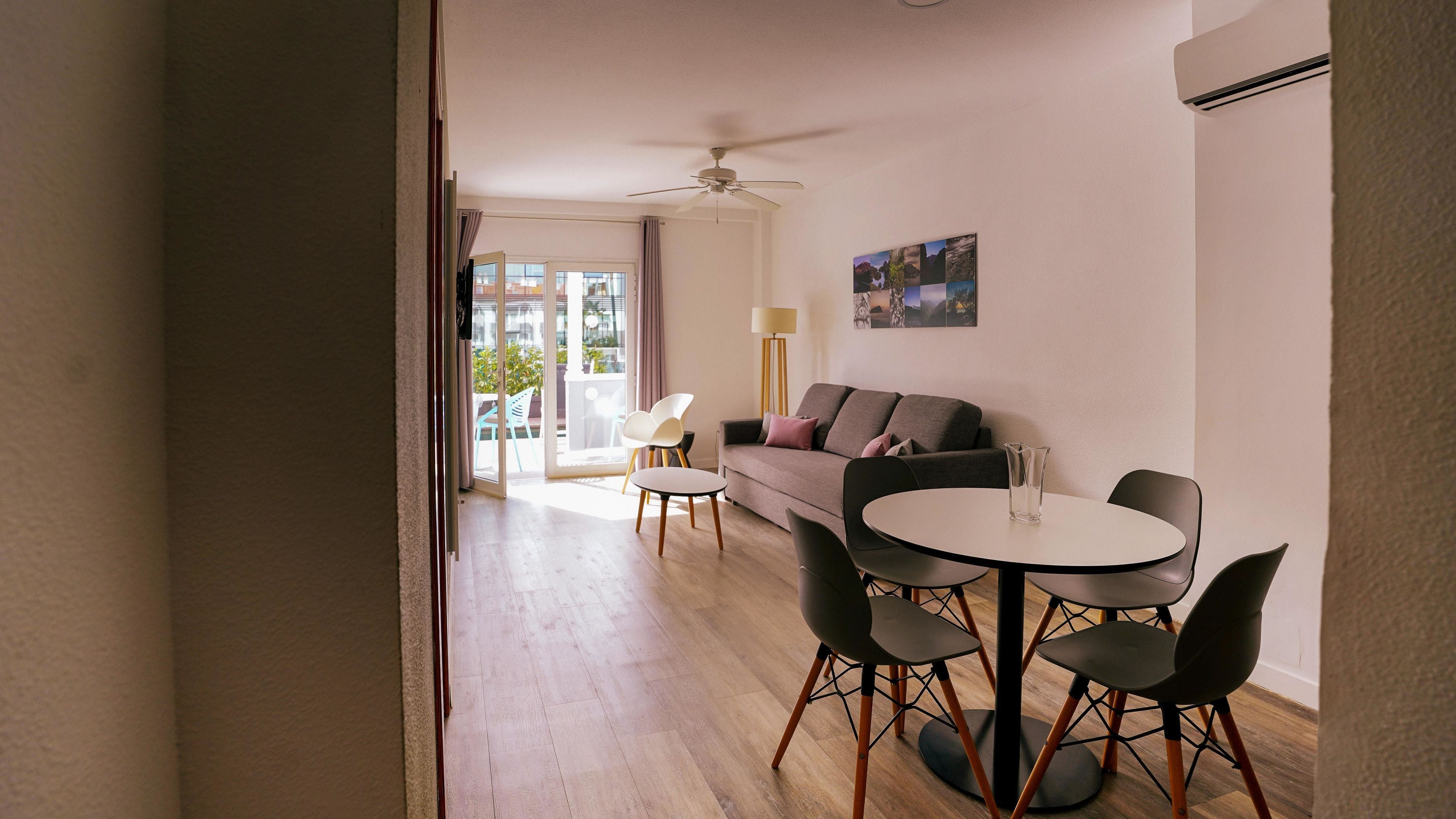 A modern living room with a sofa, a dining table with chairs, a ceiling fan, and a view through a glass door to an outdoor area.