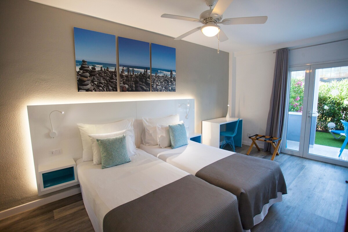 Habitación de hotel con dos camas, ventilador de techo, vistas al exterior y un balcón con mesa y sillas.