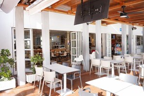 Un comedor interior con mesas y sillas blancas, un ventilador de techo y vista a la cocina al fondo.