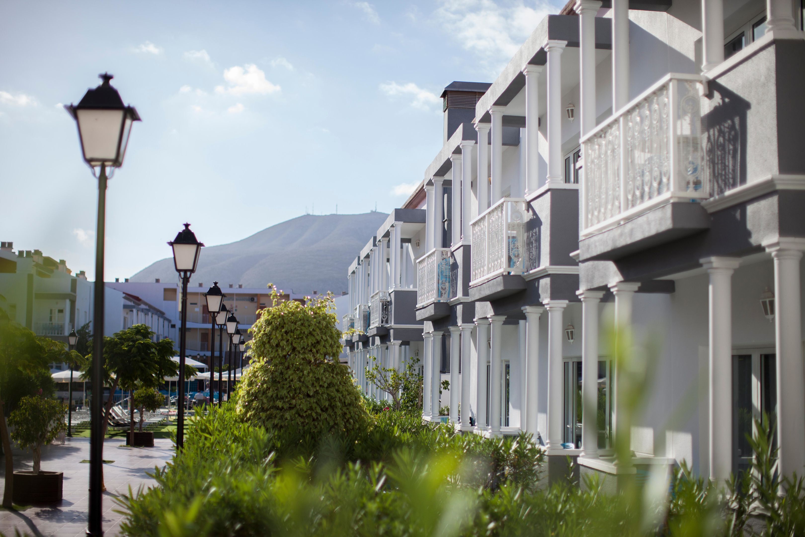 Une rangée de bâtiments blancs dotés de balcons, une rue bordée de lampadaires et une montagne en arrière-plan.